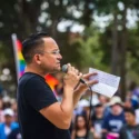 A photo of an elected official Sen. Carlos Guillermo Smith, dressed in a black t-shirt speaking to a protest crowd during a summer protest in Ybor City, Florida. There's a Pride flag in the background, and he is holding a microphone and notes.