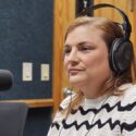 A woman in a radio studio. She wears studio monitor headphone and is listening to to the host, not in the picture, asking her a question.