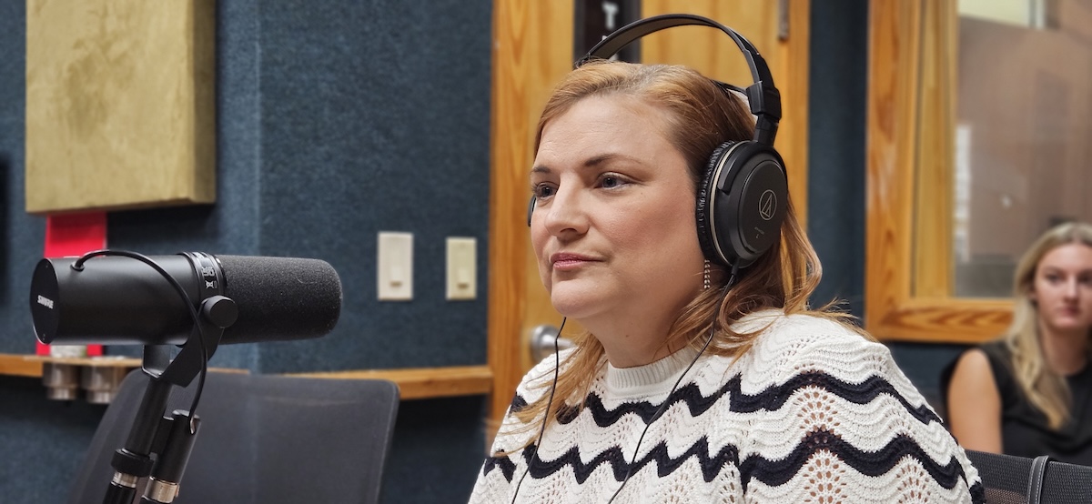 A woman in a radio studio. She wears studio monitor headphone and is listening to to the host, not in the picture, asking her a question.