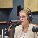 A US congresswoman, Kathy Castor, wearing a cream suit top and white shirt, with radio headphone over her head. She wears glasses and is speaking into a radio microphone.