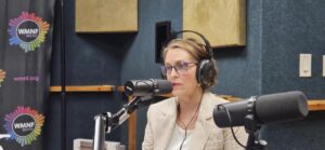 A US congresswoman, Kathy Castor, wearing a cream suit top and white shirt, with radio headphone over her head. She wears glasses and is speaking into a radio microphone.