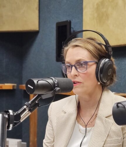 A US congresswoman, Kathy Castor, wearing a cream suit top and white shirt, with radio headphone over her head. She wears glasses and is speaking into a radio microphone.